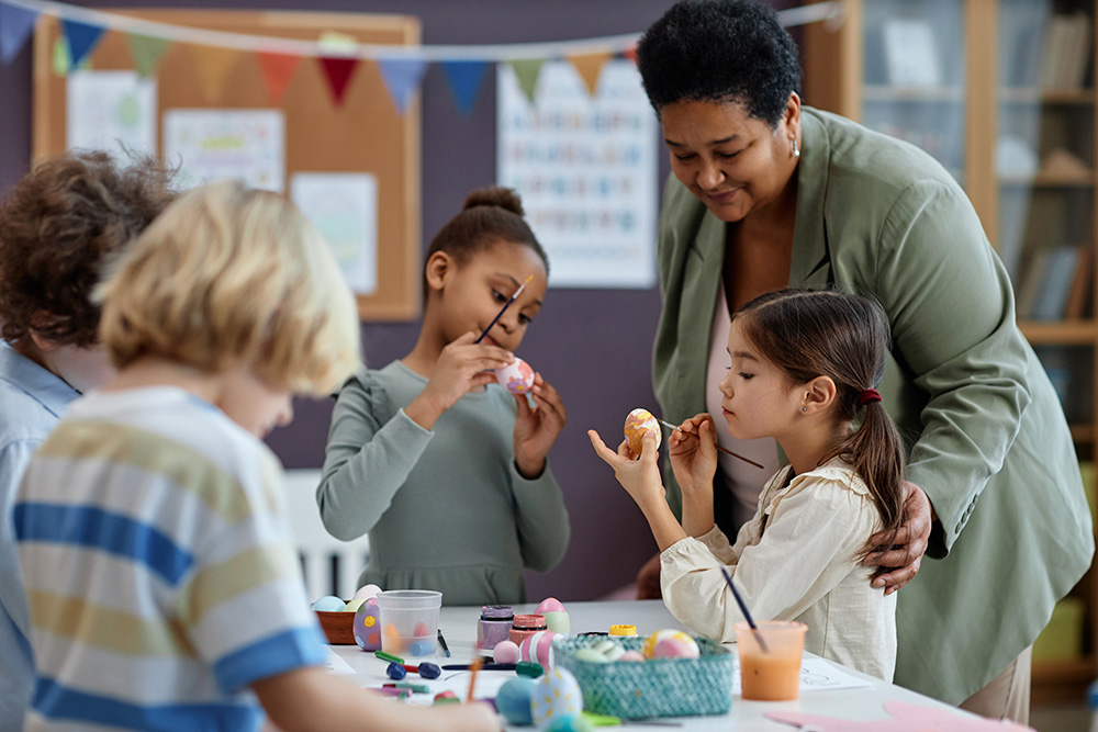 Children and a teacher collaborate at a table, creating art with a variety of colorful Easter eggs.