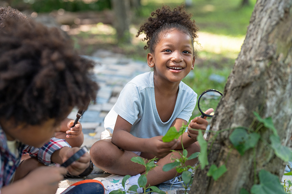 In an outdoor adventure, two children sit on the ground, captivated by the sight of a tree in their natural surroundings.