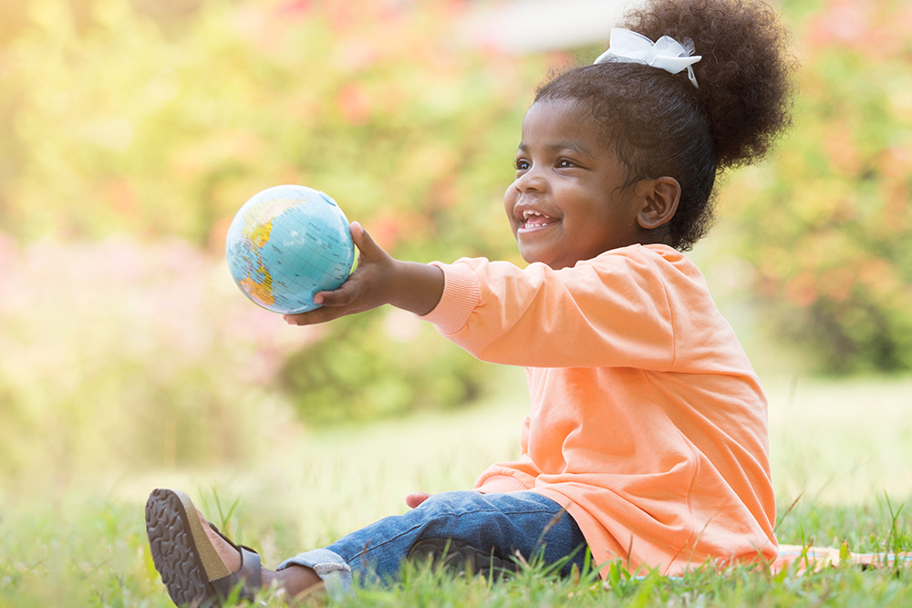 A young girl sits on the grass outdoors, joyfully holding a globe, symbolizing exploration and playfulness.