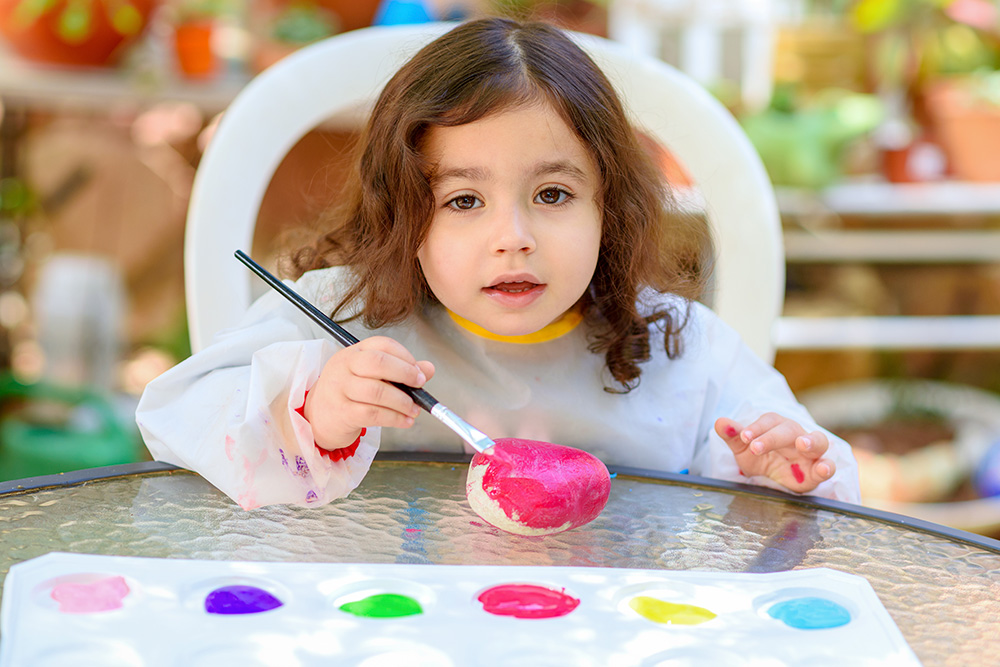 A young girl joyfully paints on a table, surrounded by colorful paints and brushes, showcasing her creativity.