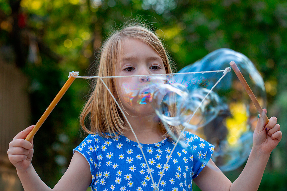 A little girl enthusiastically blows bubbles with sticks, surrounded by a playful atmosphere of floating bubbles.